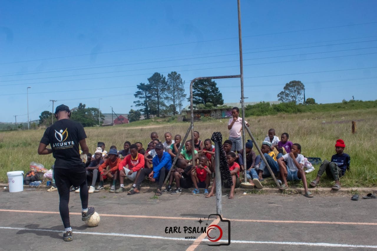A coach in a Sikunye shirt holding a soccer ball while addressing young players seated along a court